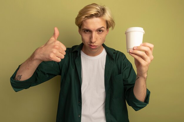 Soddisfatto giovane ragazzo biondo che indossa la maglietta verde che tiene la tazza di tè e che mostra il pollice in su