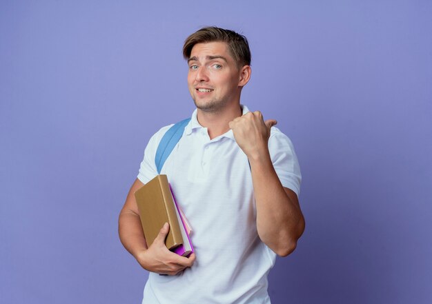 Soddisfatto giovane studente maschio bello che indossa la borsa posteriore in possesso di libri e punti lato isolato sull'azzurro