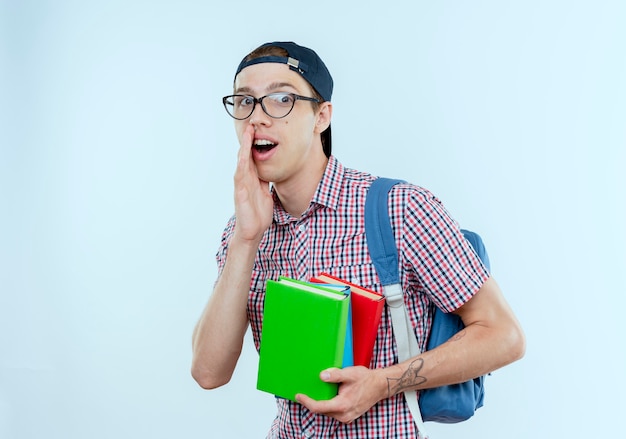 Soddisfatto giovane studente ragazzo che indossa borsa posteriore e occhiali e berretto in possesso di libri e sussurri su bianco