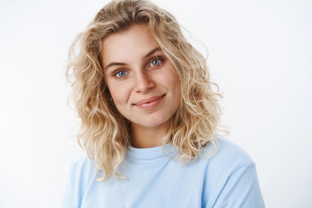 Soddisfatto, spensierato bella ragazza scandinava con gli occhi azzurri e capelli corti ricci sorridente ottimista e carina inclinando la testa civettuola mentre ascolta, guardando sincero la telecamera sul muro bianco