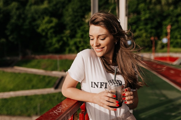 Sognando la donna felice che indossa la maglietta bianca che beve il caffè del mattino e cammina sulla strada soleggiata con un sorriso felice