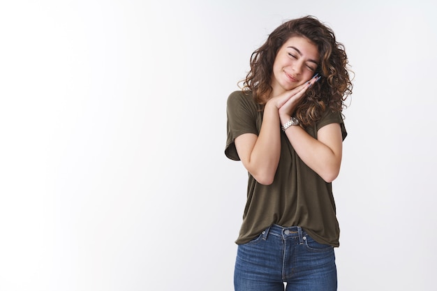 Sognante giovane donna carina magra con i palmi della testa come un cuscino che sorride e gli occhi chiusi fanno sembrare che dorma in piedi con gioia indossando jeans t-shirt verde oliva, sfondo bianco, senti il sogno che diventa realtà