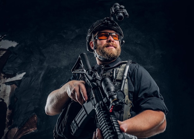 Soldato barbuto delle forze speciali che indossa un'armatura e un casco con visione notturna che tiene un fucile d'assalto. Foto dello studio contro una parete strutturata scura