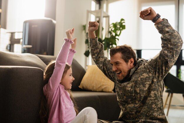 Soldato felice dell'esercito e sua piccola figlia che si divertono mentre celebrano la riunione con le braccia alzate nel soggiorno