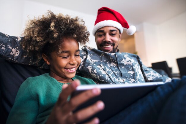 Soldato in uniforme che indossa il cappello di Babbo Natale seduto da sua figlia sul divano e utilizza il computer tablet