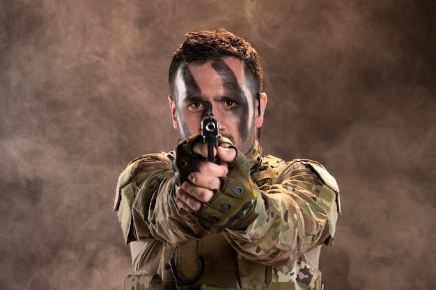 Soldato maschio in mimetica che mira pistola sul muro scuro fumoso