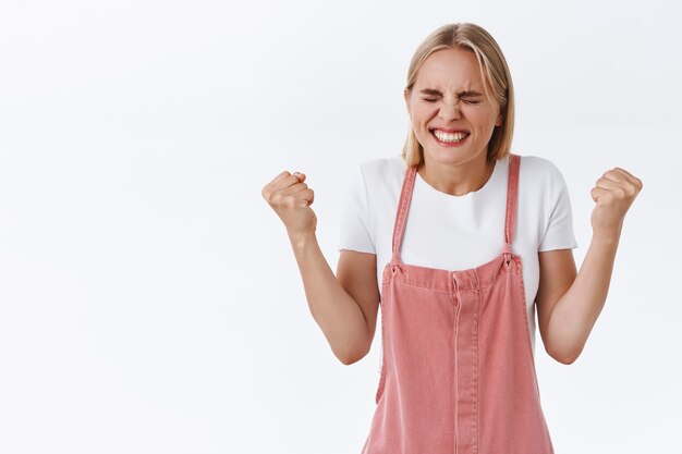 Sollevata, felice e bella giovane donna caucasica finalmente raggiunge l'obiettivo, esultando la pompa del pugno e dice di sì, strizzando gli occhi sorridendo soddisfatta, trionfando, celebrando il successo, vincendo la scommessa, sfondo bianco