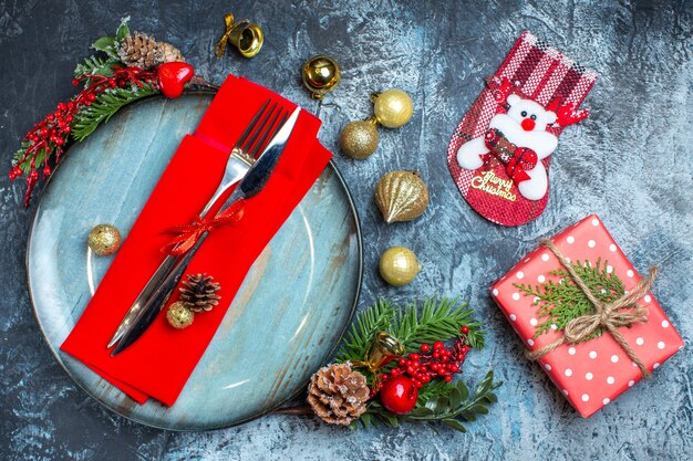 Sopra la vista del set di posate con nastro rosso su un tovagliolo decorativo su un piatto blu e accessori natalizi accanto a confezione regalo e calzino natalizio su sfondo scuro