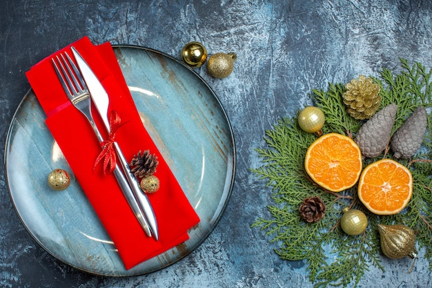 Sopra la vista del set di posate con nastro rosso su un tovagliolo decorativo su un piatto blu e collezione di accessori regalo di agrumi cono di conifere su sfondo scuro