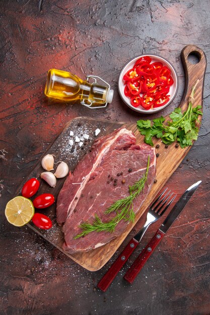 Sopra la vista della carne rossa sul tagliere di legno e della bottiglia di olio caduta del pomodoro del pepe tritato del limone verde dell'aglio su fondo scuro