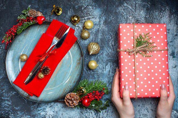 Sopra la vista della mano che tiene una confezione regalo e un set di posate con nastro rosso su un tovagliolo decorativo su un piatto blu e accessori natalizi e calzini natalizi su sfondo scuro