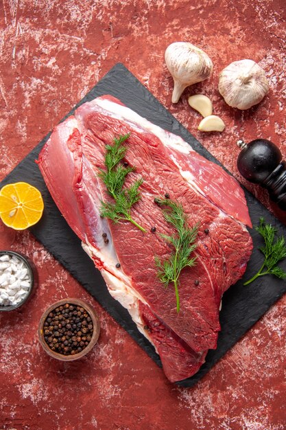 Sopra la vista di carne rossa fresca con verde e pepe su tavola nera coltello aglio limone spezie martello di legno limone su olio sfondo rosso pastello