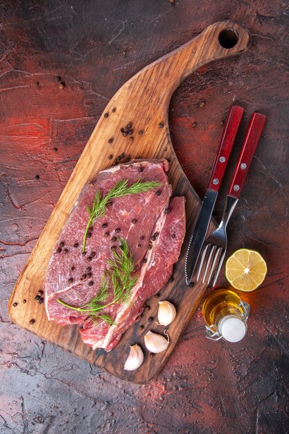 Sopra la vista di carne rossa sul tagliere di legno e forchetta e coltello della bottiglia dell'olio del pepe verde dell'aglio su fondo scuro