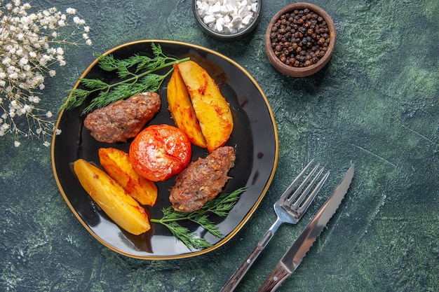 Sopra la vista di deliziose cotolette di carne al forno con patate e pomodori su un piatto nero set di posate fiori bianchi spezie su sfondo verde nero colori misti