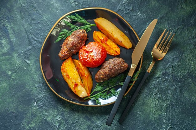 Sopra la vista di deliziose cotolette di carne al forno con patate e pomodori su una piastra nera servite con posate verdi su sfondo nero verde colori mix