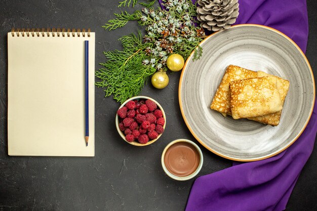 Sopra la vista di deliziose frittelle su un piatto bianco cioccolato e accessori per la decorazione di lamponi su un taccuino di asciugamani viola con penna su sfondo nero