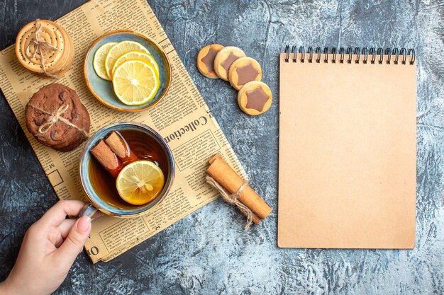 Sopra la vista di deliziosi biscotti e mano che tiene una tazza di tè nero con cannella su un vecchio giornale