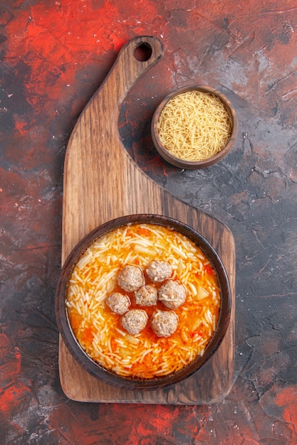 Sopra la vista di gustosa zuppa di pollo con tagliatelle su tagliere di legno e pasta cruda in una piccola ciotola su sfondo scuro