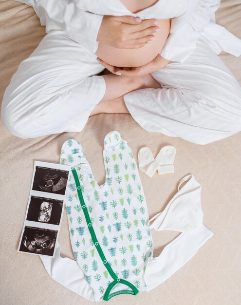 Sopra la vista di una donna anonima in un accogliente pigiama bianco seduta sul letto con l'immagine ad ultrasuoni di un grazioso body e calzini dell'utero mentre accarezza la pancia incinta e aspetta il bambino e parte della famiglia