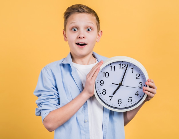 Sorprenda il ragazzo sorridente che tiene l&#39;orologio bianco a disposizione che guarda alla macchina fotografica contro fondo giallo