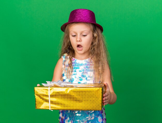 sorpresa bimba bionda con cappello da festa viola che tiene in mano e guarda la confezione regalo isolata sulla parete verde con spazio copia