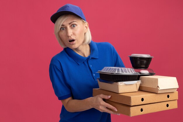 sorpresa bionda di mezza età donna di consegna in uniforme blu e berretto che tiene pacchetti di pizza con contenitori per alimenti e pacchetto di cibo di carta su di loro guardando davanti