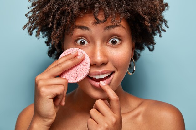 Sorpresa e allegra donna afroamericana sente consigli su come prendersi cura della pelle, tiene la spugna cosmetica sulla guancia, ha gli occhi ampiamente aperti, reazione scioccata, rimuove il trucco. Spa e concetto di relax