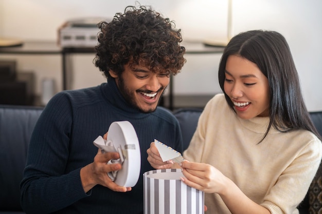 Sorpresa, gioia. Sorridente attento uomo indiano dai capelli ricci che apre la scatola luminosa del regalo e bella donna asiatica piacevolmente sorpresa che estrae la lettera seduta sul divano