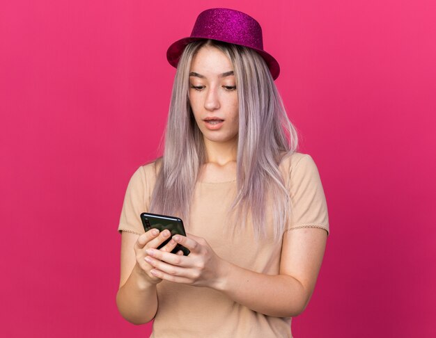Sorpresa giovane bella donna che indossa un cappello da festa che tiene e guarda il telefono isolato sul muro rosa