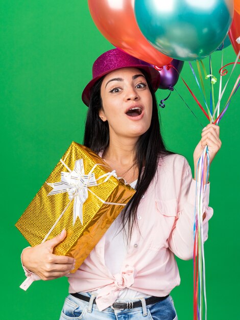 Sorpresa giovane bella donna che indossa un cappello da festa con palloncini che tengono una scatola regalo isolata sul muro verde