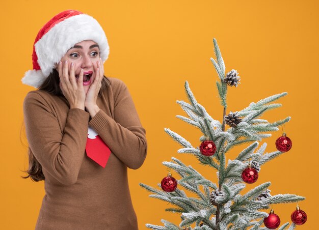 sorpresa giovane bella ragazza che indossa il cappello di natale con cravatta in piedi vicino albero di natale mettendo le mani sulle guance isolate sulla parete arancione