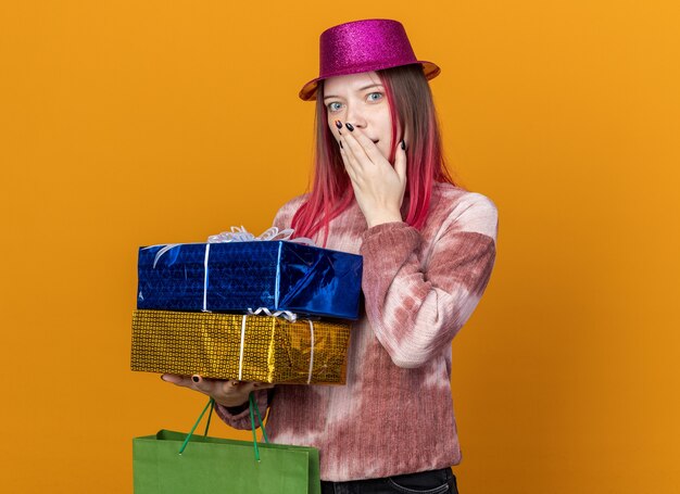 Sorpresa giovane bella ragazza che indossa un cappello da festa che tiene in mano una borsa regalo con scatole regalo coprendo il viso con la mano