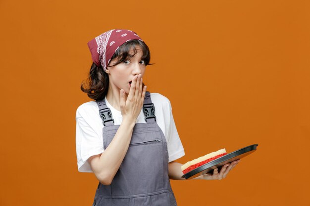 Sorpresa giovane donna addetta alle pulizie che indossa uniforme e bandana vassoio con spugna in esso guardando la fotocamera tenendo la mano sulla bocca isolata su sfondo arancione