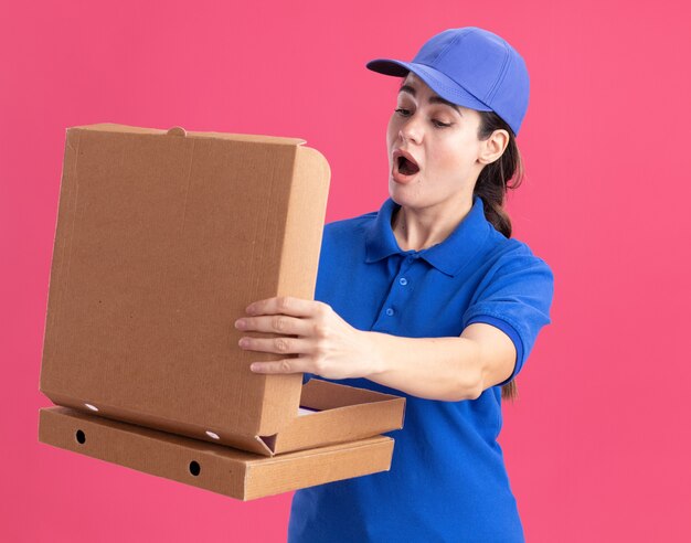 Sorpresa giovane donna delle consegne in uniforme e berretto che tiene in mano i pacchetti di pizza aprendone uno che ci guarda dentro