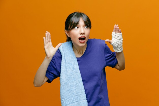 Sorpresa giovane donna sportiva che indossa una maglietta guardando il lato che mostra le mani vuote con un asciugamano sulla spalla con il polso avvolto con un bendaggio isolato su sfondo arancione