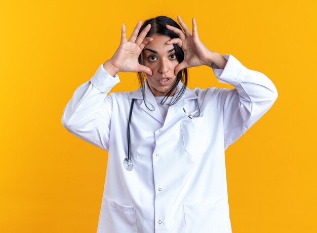 Sorpresa giovane dottoressa che indossa un abito medico con uno stetoscopio che apre gli occhi con le mani isolate su sfondo giallo