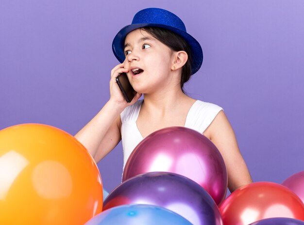 sorpresa giovane ragazza caucasica che indossa il cappello blu del partito parlando al telefono guardando il lato in piedi con palloncini di elio isolato sul muro viola con spazio di copia