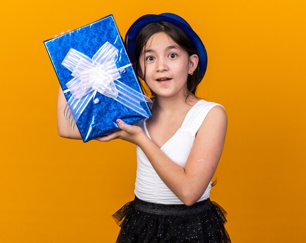 sorpresa giovane ragazza caucasica con blue party hat holding confezione regalo isolata sulla parete arancione con copia space