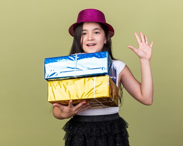 sorpresa giovane ragazza caucasica con cappello da festa viola che tiene scatole regalo e in piedi con la mano alzata isolata sul muro verde oliva con spazio copia