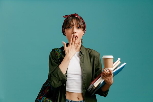 Sorpresa giovane studentessa che indossa bandana e zaino con in mano una tazza di caffè di carta e blocchi per appunti guardando la fotocamera facendo un gesto oops isolato su sfondo blu