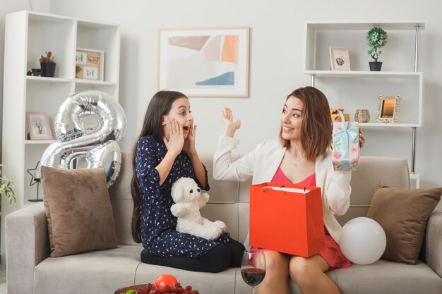 Sorpreso a guardarsi l'un l'altro bambina e madre con regalo e orsacchiotto in felice festa della donna seduta sul divano in soggiorno