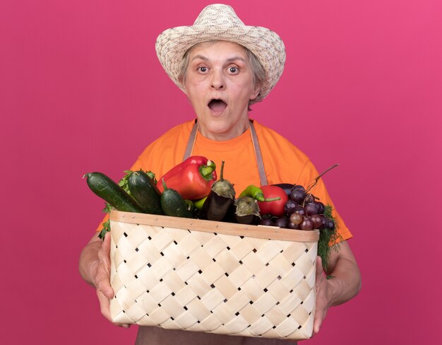 Sorpreso anziano giardiniere femminile che indossa cappello da giardinaggio che tiene cesto di verdure vegetable