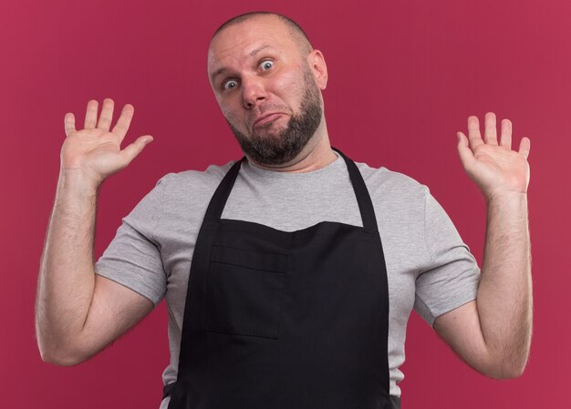 Sorpreso barbiere maschio di mezza età in uniforme che allarga le mani isolate sulla parete rosa