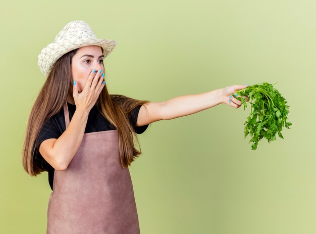 Sorpreso bella ragazza giardiniere in uniforme che indossa cappello da giardinaggio tenendo fuori il coriandolo a lato e la bocca coperta con la mano isolata su verde oliva