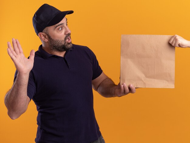 Sorpreso fattorino di mezza età in uniforme e berretto che dà un pacchetto di cibo di carta al cliente isolato sul muro giallo