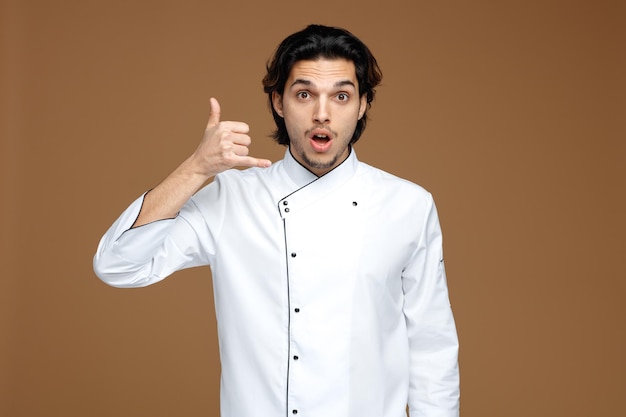 sorpreso giovane chef maschio che indossa l'uniforme guardando la fotocamera che mostra il gesto di chiamata isolato su sfondo marrone