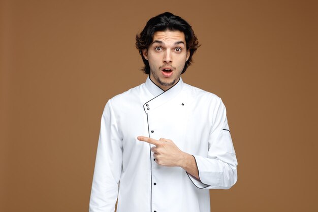 sorpreso giovane chef maschio che indossa l'uniforme guardando la fotocamera che punta al lato isolato su sfondo marrone