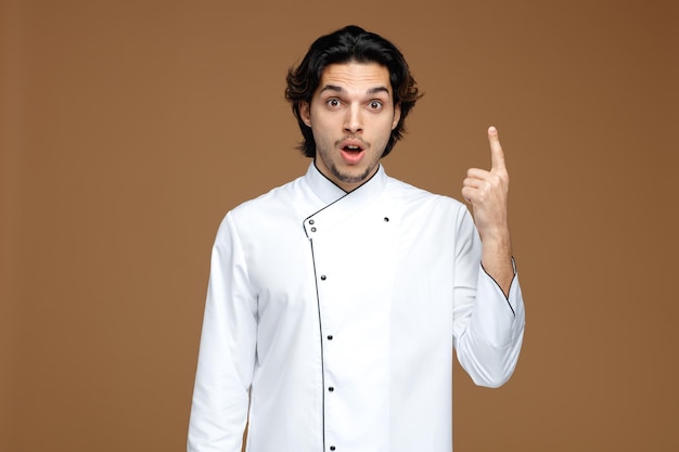 sorpreso giovane chef maschio che indossa l'uniforme guardando la fotocamera rivolta verso l'alto isolata su sfondo marrone