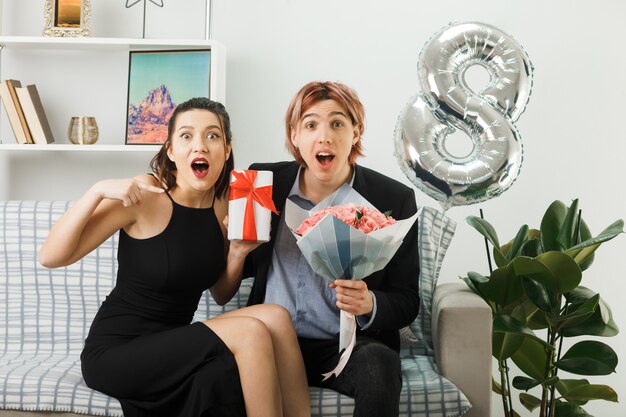 Sorpreso giovane coppia il giorno delle donne felici tenendo presente con bouquet seduto sul divano in soggiorno