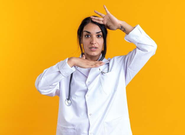 Sorpreso giovane dottoressa che indossa un abito medico con uno stetoscopio che si tiene per mano intorno al viso isolato su sfondo giallo